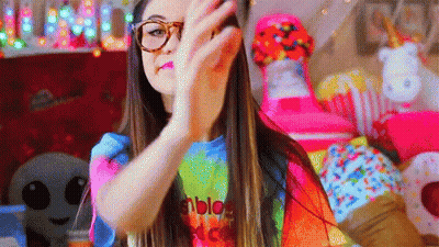 A Girl Wearing Glasses And A Tie Dye Shirt Is Making A Peace Sign . GIF