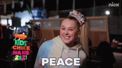 A Girl With A Bow In Her Hair Is Smiling In Front Of A Sign That Says Kids Choice Awards 2021 GIF