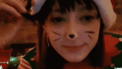 A Girl With A Cat Face Painted On Her Face And A Santa Hat GIF