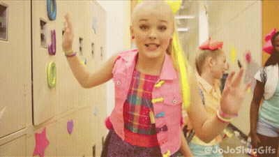 A Girl With A Yellow Bow In Her Hair Is Standing In A Hallway With Other Girls GIF