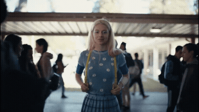 A Girl With Blonde Hair And A Blue Shirt Is Standing In A Hallway GIF