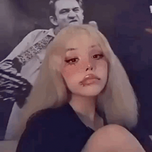 A Girl With Blonde Hair And Freckles Is Sitting In Front Of A Picture Of A Man Playing A Guitar . GIF