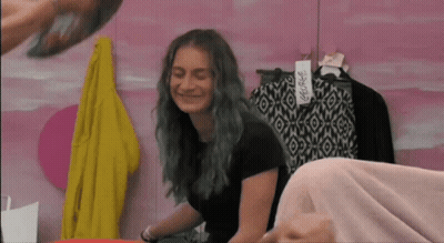 A Girl With Blue Hair Is Smiling In Front Of A Pink Wall With Clothes Hanging On It GIF