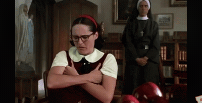 A Girl With Glasses And A Red Headband Is Sitting At A Table With Her Arms Crossed In Front Of A Nun . GIF