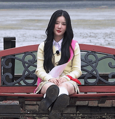 A Girl With Long Black Hair Is Sitting On A Bench With Her Legs Crossed GIF