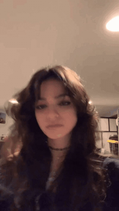 A Girl With Long Dark Hair Is Standing In A Room . GIF