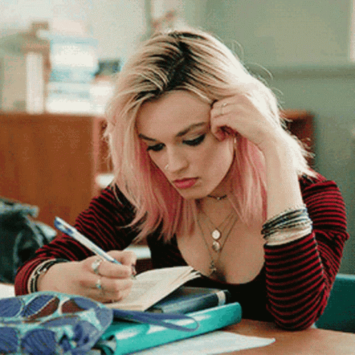 A Girl With Pink Hair Is Sitting At A Desk Writing In A Notebook . GIF