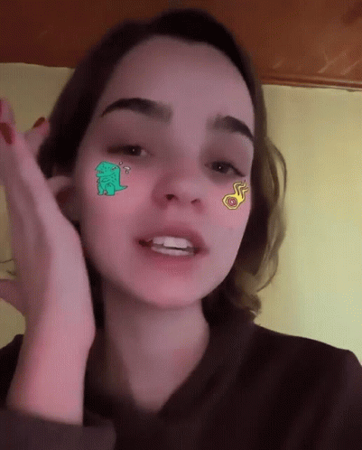 A Girl With Stickers On Her Face Including A Green Dinosaur GIF