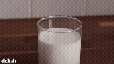 A Glass Of Milk Is Sitting On A Wooden Table With The Word Delish On The Bottom . GIF