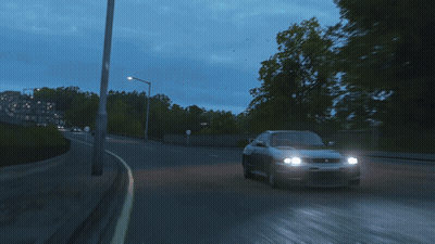 A Gray Sports Car Is Driving Down A Road With Trees In The Background GIF