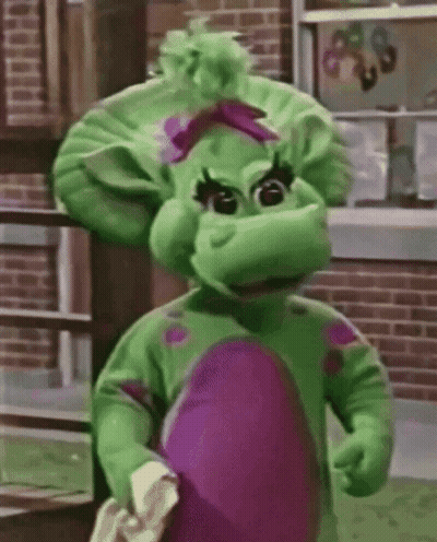 A Green And Purple Dinosaur Mascot Standing In Front Of A Brick Building . GIF