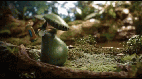 A Green Frog With A Mushroom On Its Head Is Standing In A Swamp GIF