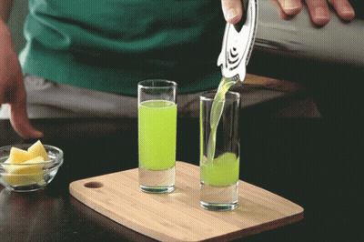 A Green Liquid Is Being Poured Into Two Shot Glasses On A Cutting Board GIF