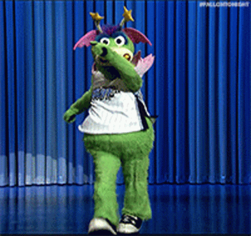 A Green Mascot Is Dancing On A Stage With A Blue Curtain Behind Him GIF
