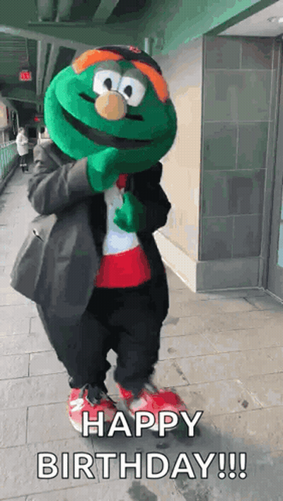A Green Mascot Is Standing On A Sidewalk And Says Happy Birthday ! GIF