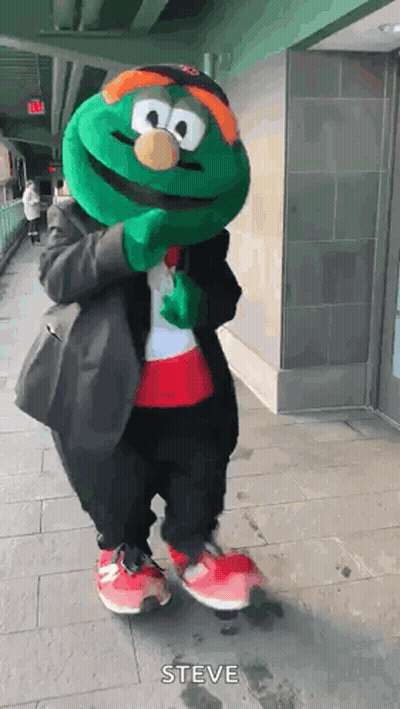 A Green Mascot Is Standing On A Sidewalk Wearing A Suit And A Hat . GIF
