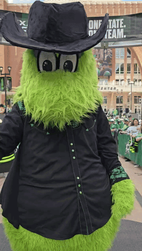 A Green Mascot Is Wearing A Black Cowboy Hat And A Black Shirt GIF