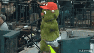 A Green Mascot Wearing A Red Hat And Shorts Is Dancing In The Stands At A Baseball Game .. GIF