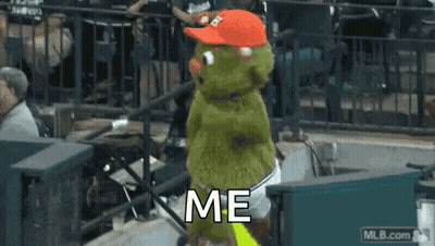 A Green Stuffed Animal Wearing A Red Hat Is Standing In A Stadium . GIF