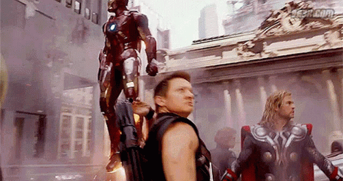 A Group Of Avengers Are Standing In Front Of A Building With A Statue Of Iron Man In The Background . GIF