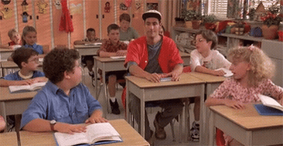 A Group Of Children Are Sitting At Their Desks In A Classroom With A Man In A Red Shirt Standing In The Middle . GIF