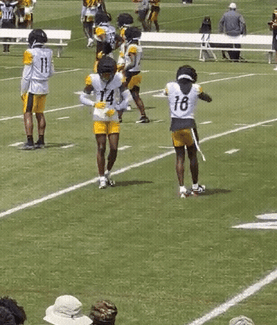 A Group Of Football Players Are Practicing On A Field While A Man In A Yellow Shirt Watches . GIF