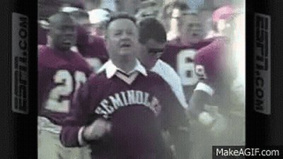 A Group Of Football Players Are Standing Around A Man Wearing A Sweater That Says ' Miami ' On It . GIF