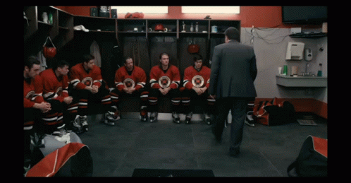 A Group Of Hockey Players Sit In A Locker Room With A Man In A Suit Talking To Them GIF
