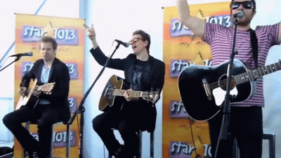 A Group Of Men Are Singing And Playing Guitars In Front Of A Banner That Says Star 1013 GIF
