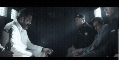 A Group Of Men Are Sitting In A Dark Room Talking To Each Other . One Of The Men Is Holding A Gun . GIF