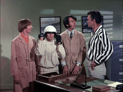 A Group Of Men Are Standing Around A Desk And One Of Them Is Wearing A White Helmet GIF
