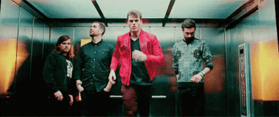 A Group Of Men Are Standing In An Elevator . One Of The Men Is Wearing A Red Jacket . GIF