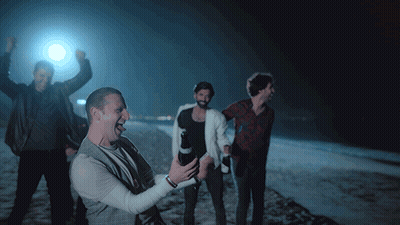 A Group Of Men Are Standing On A Beach At Night And One Of Them Is Holding A Bottle Of Beer GIF