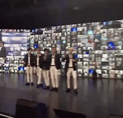 A Group Of Men Are Standing On A Stage In Front Of A Wall Of Television Screens GIF