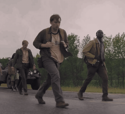 A Group Of Men Are Walking Down A Road And One Of Them Has A Badge On His Chest GIF