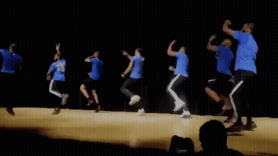 A Group Of Men In Blue Shirts Are Dancing On A Stage In Front Of A Crowd GIF