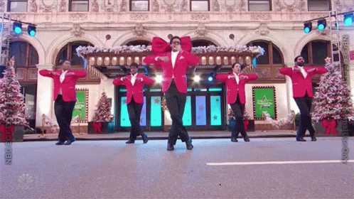 A Group Of Men In Red Jackets Are Dancing In Front Of A Building With Christmas Trees GIF