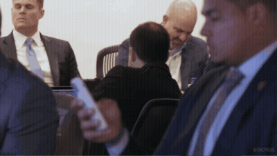 A Group Of Men In Suits And Ties Are Sitting Around A Table And One Man Is Holding A Cell Phone In His Hand GIF