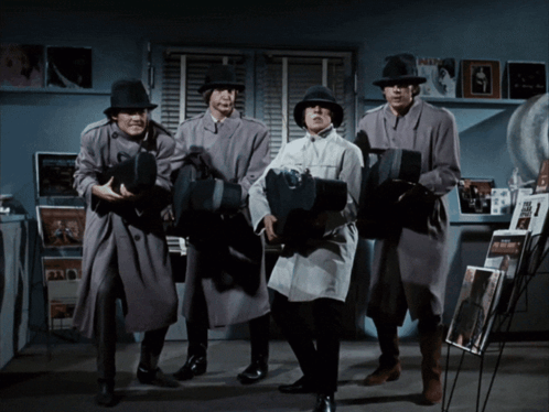 A Group Of Men In Trench Coats And Hats Are Standing In A Room Holding Cameras GIF