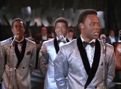A Group Of Men In Tuxedos Are Singing Into Microphones . GIF
