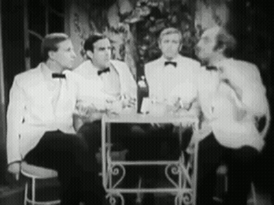 A Group Of Men In Tuxedos Are Sitting At A Table Talking To Each Other . GIF