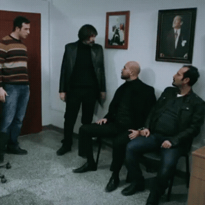 A Group Of Men Standing And Sitting In A Room With A Picture On The Wall GIF