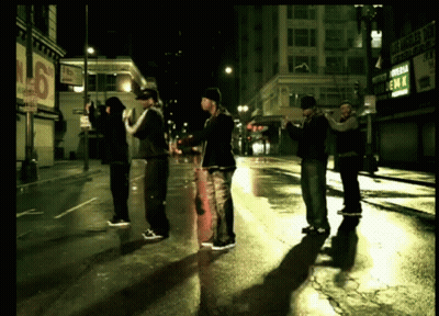 A Group Of Men Standing On A Wet Street In Front Of A Sign That Says Emm GIF