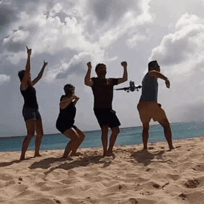 A Group Of People Are Dancing On A Beach While A Plane Is Flying In The Background . GIF