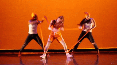 A Group Of People Are Dancing On A Stage And One Of Them Is Wearing An Orange Outfit With The Number 12 On It GIF
