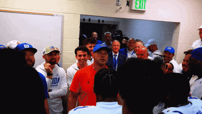 A Group Of People Are Gathered In A Room With A Green Exit Sign Above Them GIF