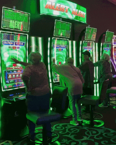 A Group Of People Are Playing A Slot Machine Under A Green Sign That Says Alert Alert GIF