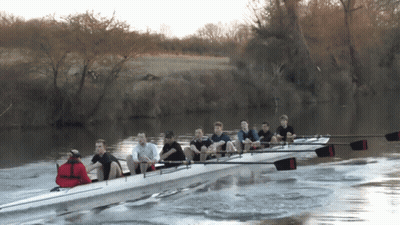 A Group Of People Are Rowing A Boat On A River With Trees In The Background GIF