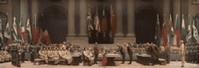 A Group Of People Are Sitting Around A Table In A Large Room With Flags Behind Them . GIF