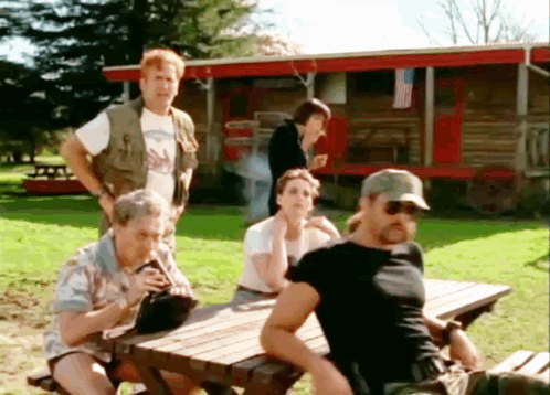 A Group Of People Are Sitting At A Picnic Table In Front Of A Cabin GIF
